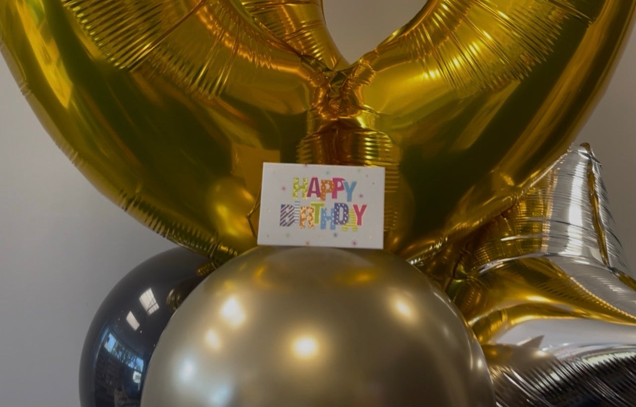 A close-up of a balloon bouquet with metallic gold foil balloons, a large gold balloon arching overhead, a silver foil balloon on the right, a metallic gold sphere at the bottom center, and a small white card reading “HAPPY BIRTHDAY” in colorful letters.