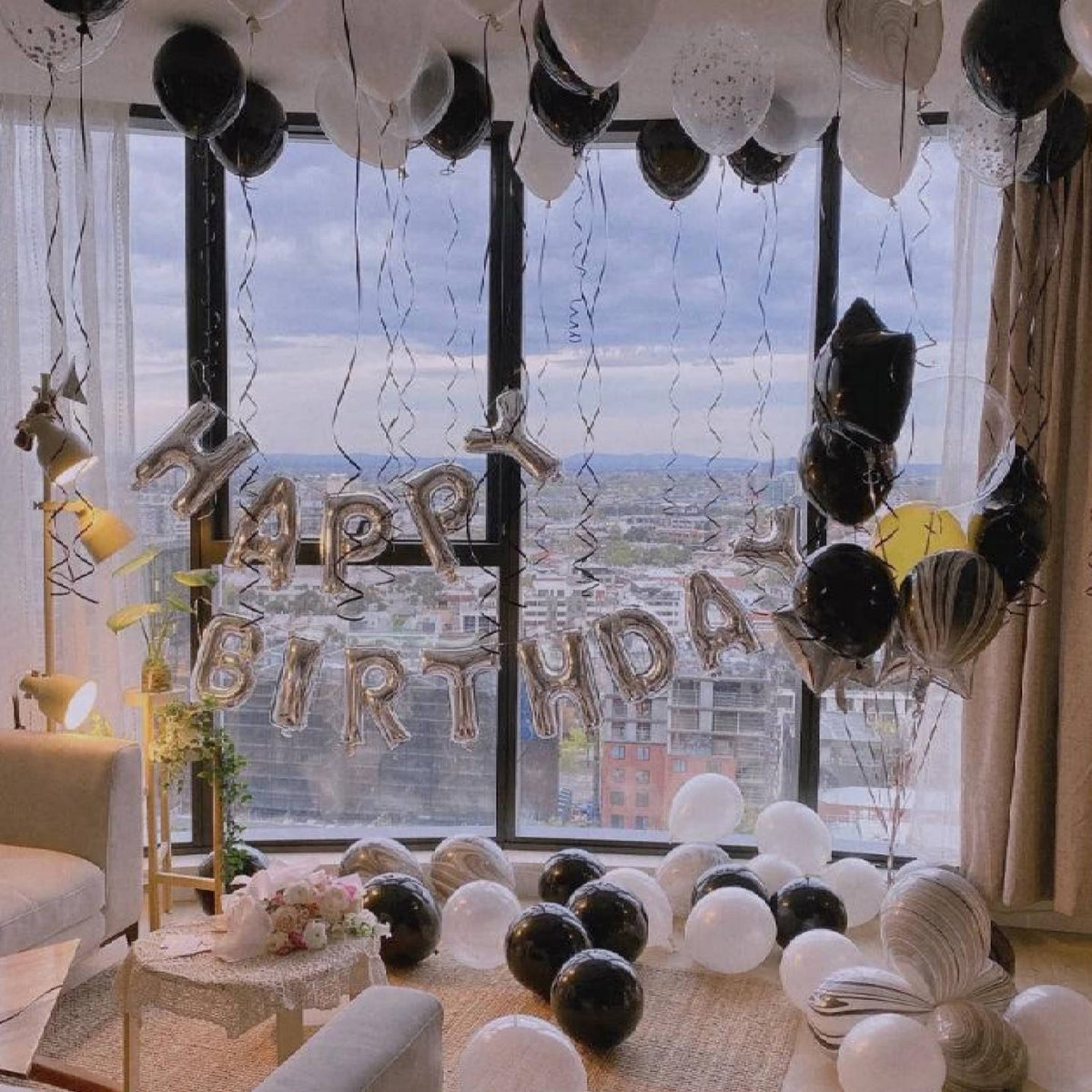 Birthday celebration with balloons and 'Happy Birthday' sign in a room with large windows.