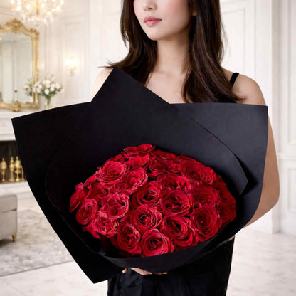 Woman holding a large bouquet of red roses wrapped in black paper indoors.