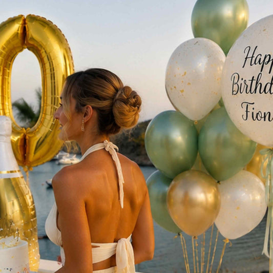 A woman seen from behind wearing a white halter bikini top, standing outdoors by the water with large gold and white balloons, including a gold number 0 balloon and a white balloon that reads 'Happy Birthday Fiona'.