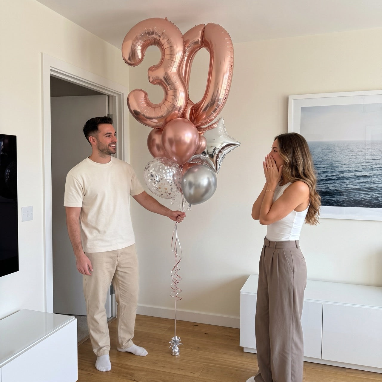 A man and a woman standing in a bright living room with wooden floors, holding a large rose gold foil balloon shaped like the number 30 with smaller silver, white, and clear balloons.