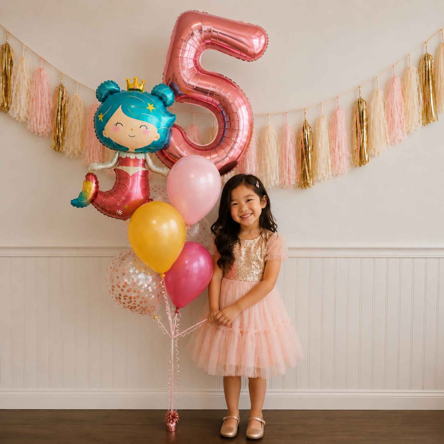 A young girl in a pink party dress stands next to a balloon bouquet featuring a large metallic pink number 5 balloon and a mermaid-themed foil balloon, set against a decorated party backdrop with tinsel garlands.