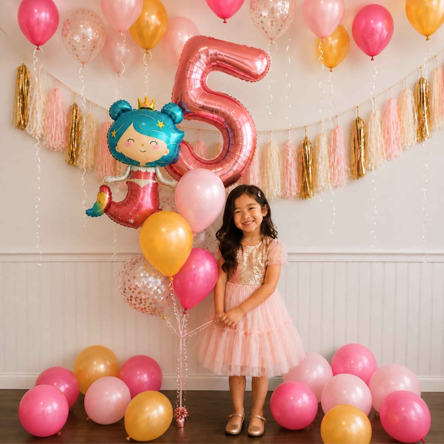 A young girl in a pink tulle dress stands in a decorated party room with pink, gold, and peach balloons, a large metallic pink number 5 balloon, a cartoon princess balloon, and gold and pink tassel garlands on the wall.