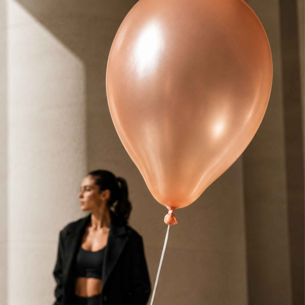 A large metallic rose gold balloon floating in the foreground, with a woman in a black blazer and crop top blurred in the background against a warm beige wall.