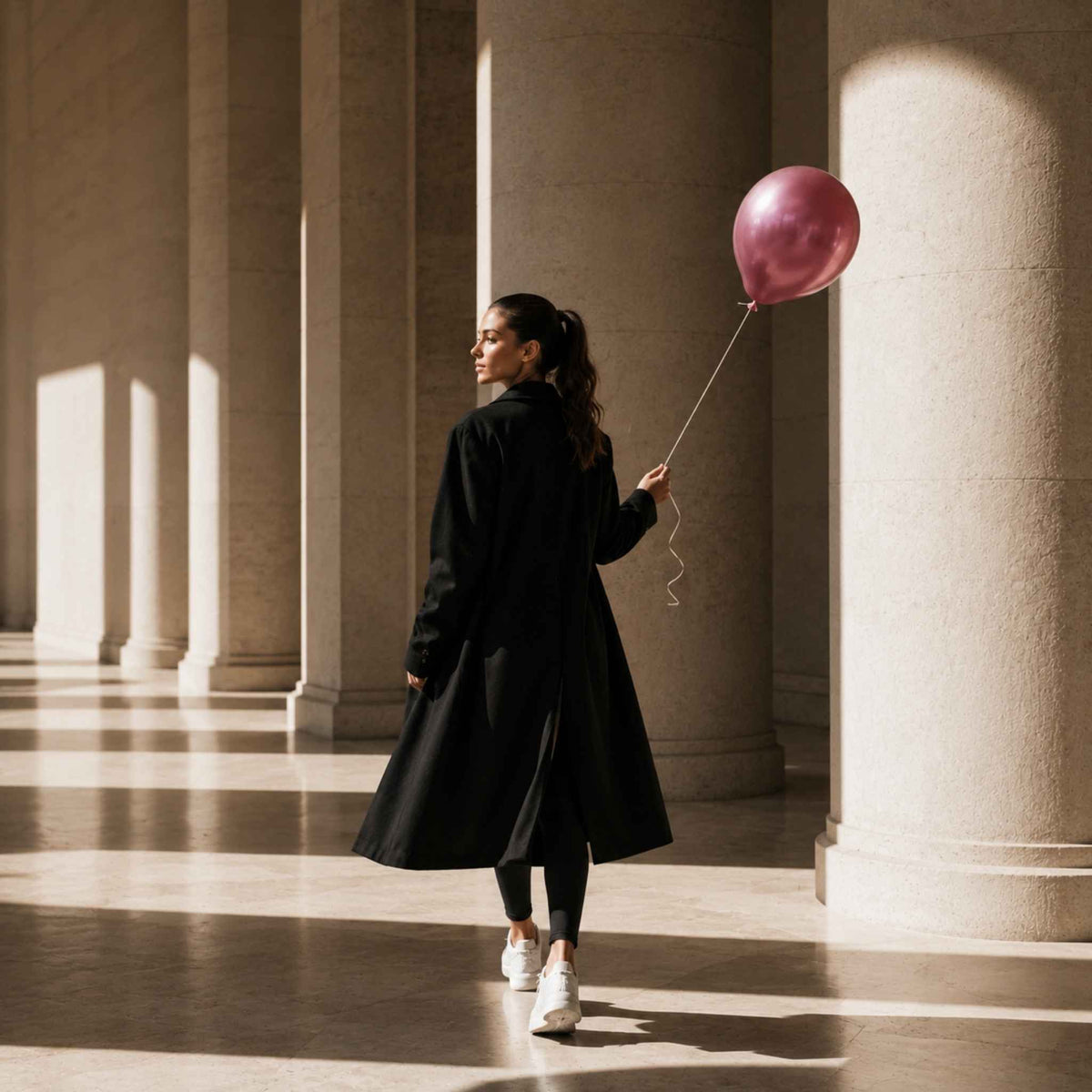 A woman in a long black coat walks through a grand hall with large stone columns, holding a metallic pink balloon. She is wearing black leggings and white sneakers.