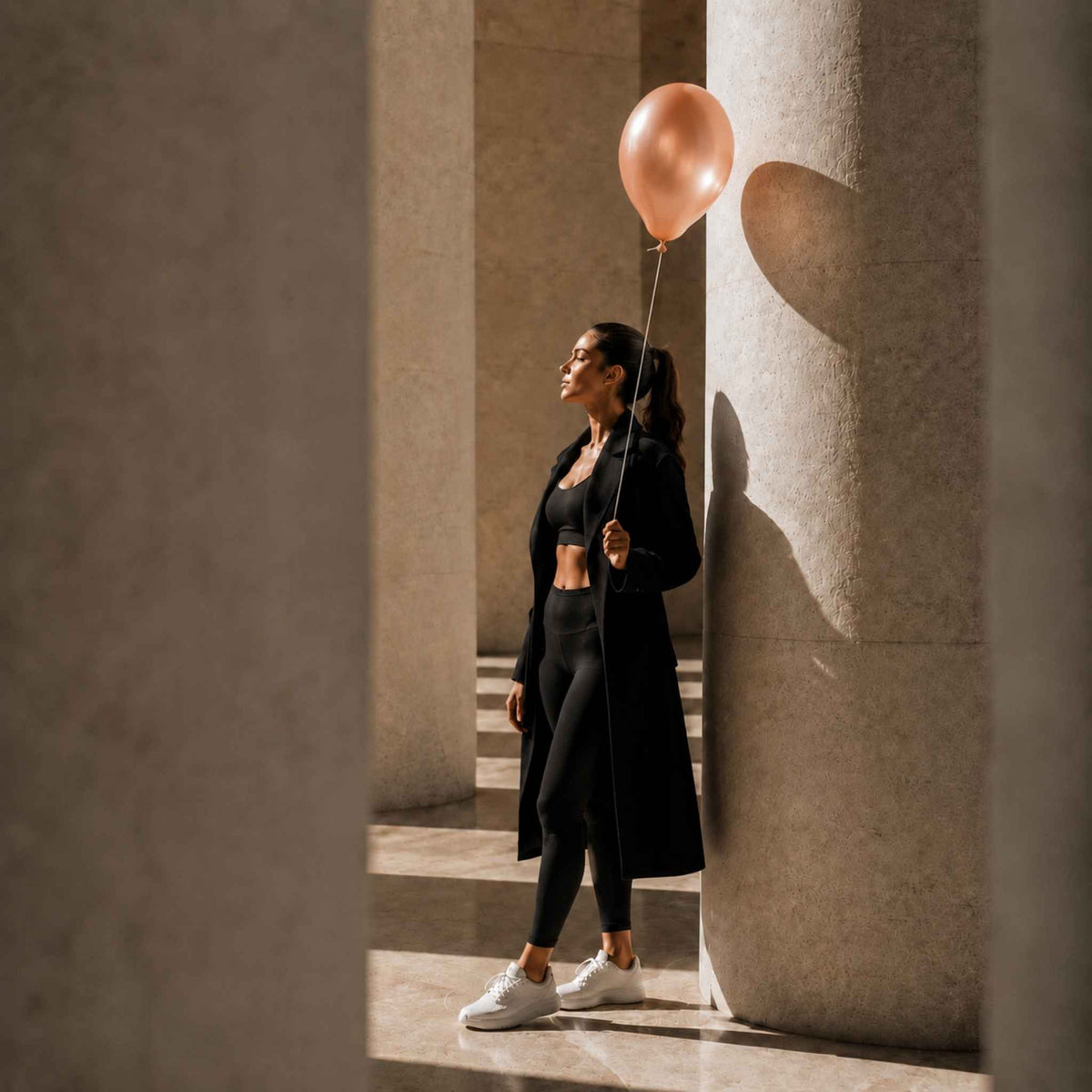 A woman in a black activewear set and long black coat stands between large stone columns, holding a metallic rose gold balloon. She is wearing white sneakers in a sunlit architectural setting.
