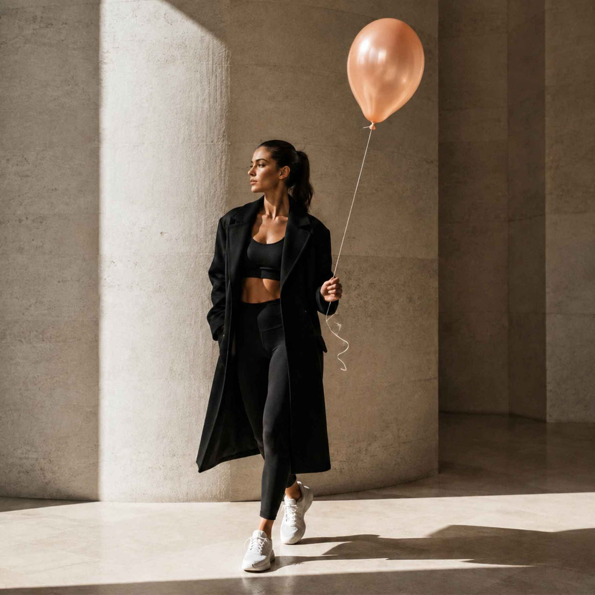 A woman in a black athletic outfit and long black coat walks through a sunlit concrete corridor while holding a metallic rose-gold balloon.