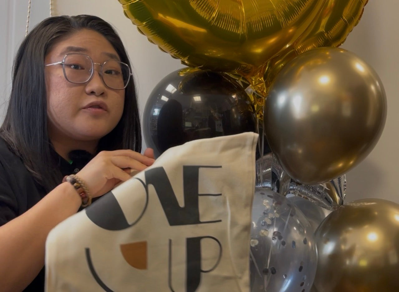 A woman wearing glasses holds a cream-colored fabric banner with the text 'ONE UP' in black and a small orange accent. The background features a cluster of metallic gold and black balloons.