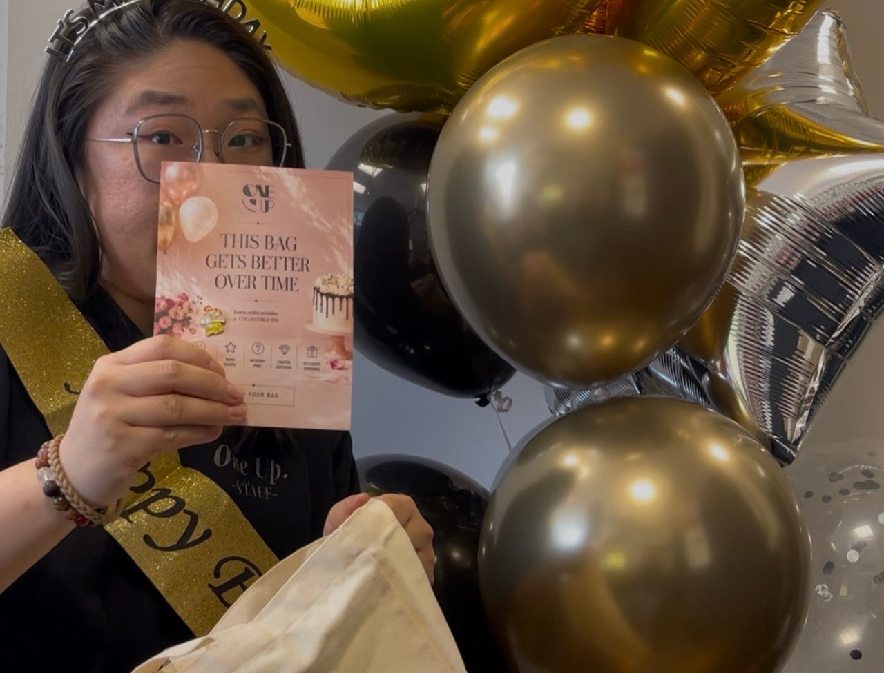 A woman wearing a gold sash holds up a pink promotional card that reads 'THIS BAG GETS BETTER OVER TIME' while surrounded by gold and black balloons.