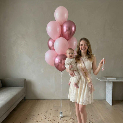 Woman holding a baby and pink balloons in a room with a couch and table.