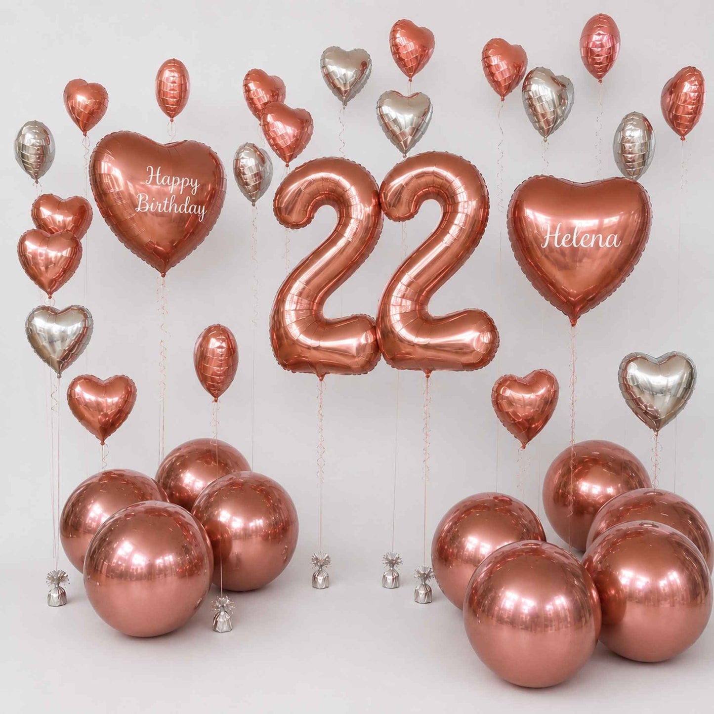 A birthday balloon set with metallic rose gold and silver foil balloons, including two large number 22 balloons, two heart balloons reading 'Happy Birthday' and 'Helena', and several round balloons clustered at the bottom on a light gray background.