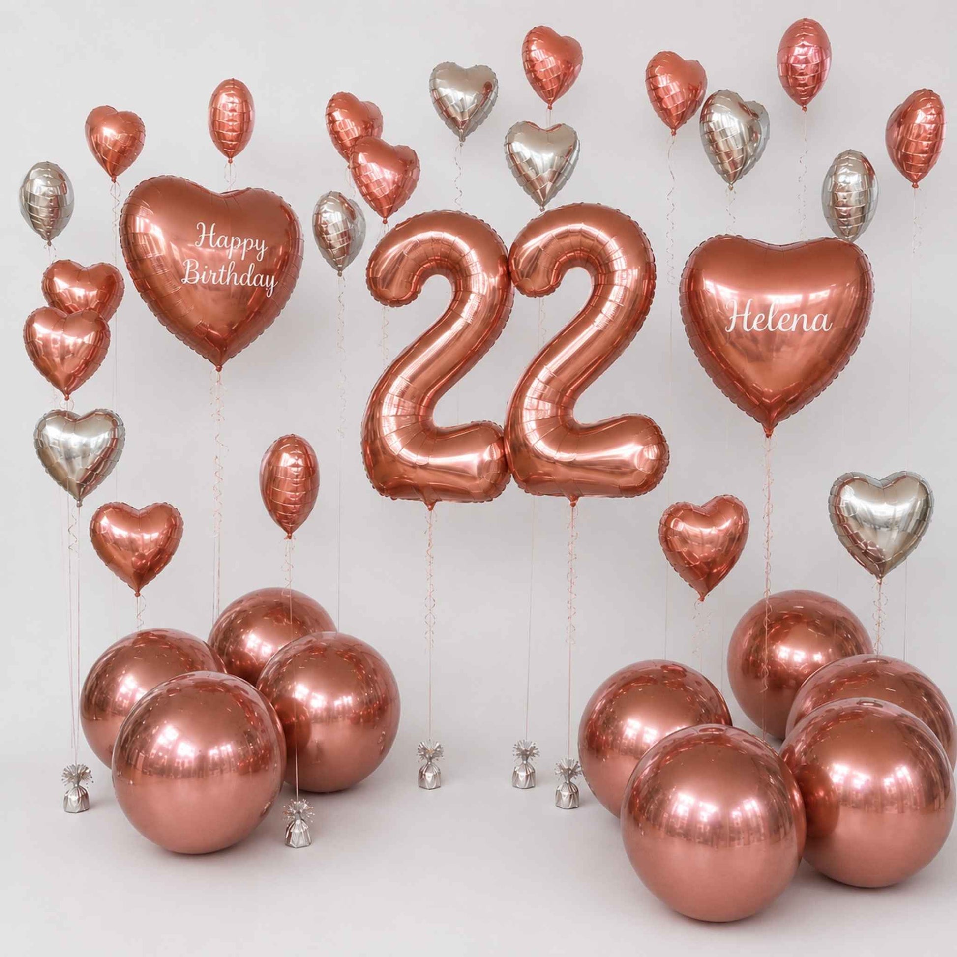 A birthday balloon set with metallic rose gold and silver foil balloons, including two large number 22 balloons, two heart balloons reading 'Happy Birthday' and 'Helena', and several round balloons clustered at the bottom on a light gray background.