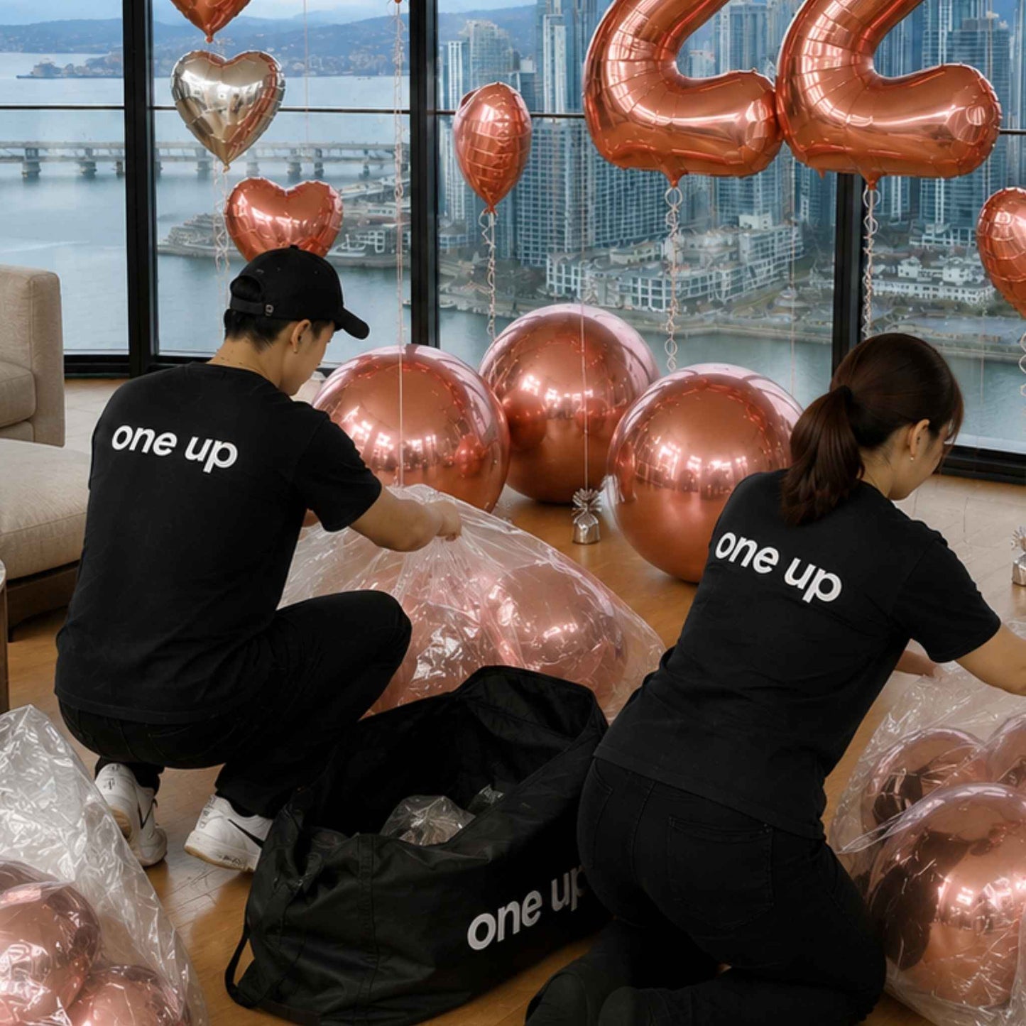Two people in black t-shirts with 'one up' printed on the back are unpacking a large set of metallic rose gold balloons, including large round balloons, heart balloons, and number balloons, from a black carrying bag in a room with a city waterfront view.