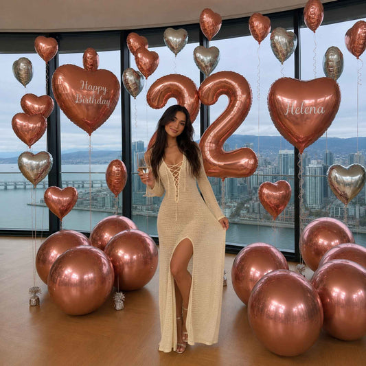 A woman in a cream lace-up maxi dress stands in a room decorated with metallic rose gold and silver balloons, including a large number 2 balloon, a 'Happy Birthday' balloon, and a heart balloon reading 'Helena'.