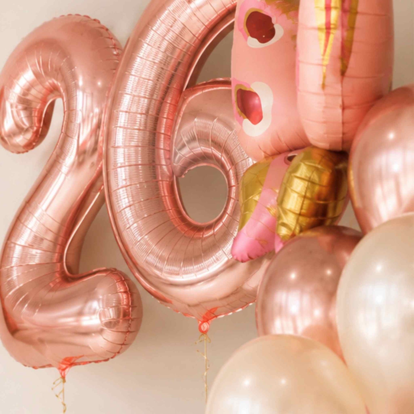 Rose gold metallic foil balloons arranged to spell the number 26, alongside a pink cartoon character foil balloon and several pastel balloons in a soft studio setting.