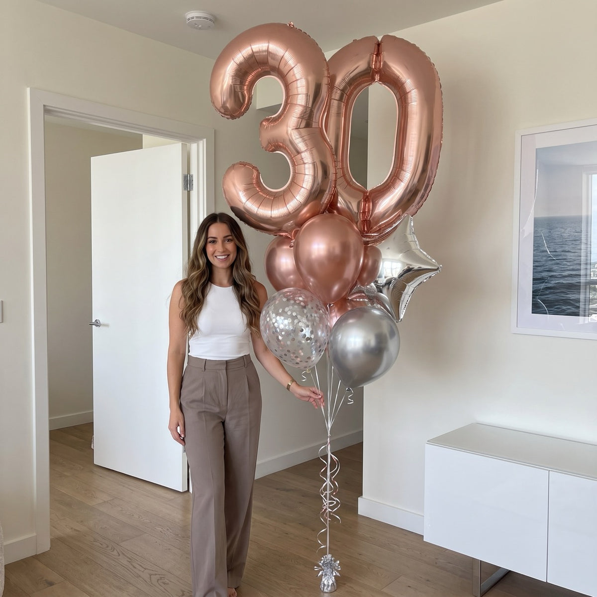 A woman stands indoors next to a large balloon bouquet featuring metallic rose gold and silver balloons, including two large number 30 balloons and a star-shaped balloon.