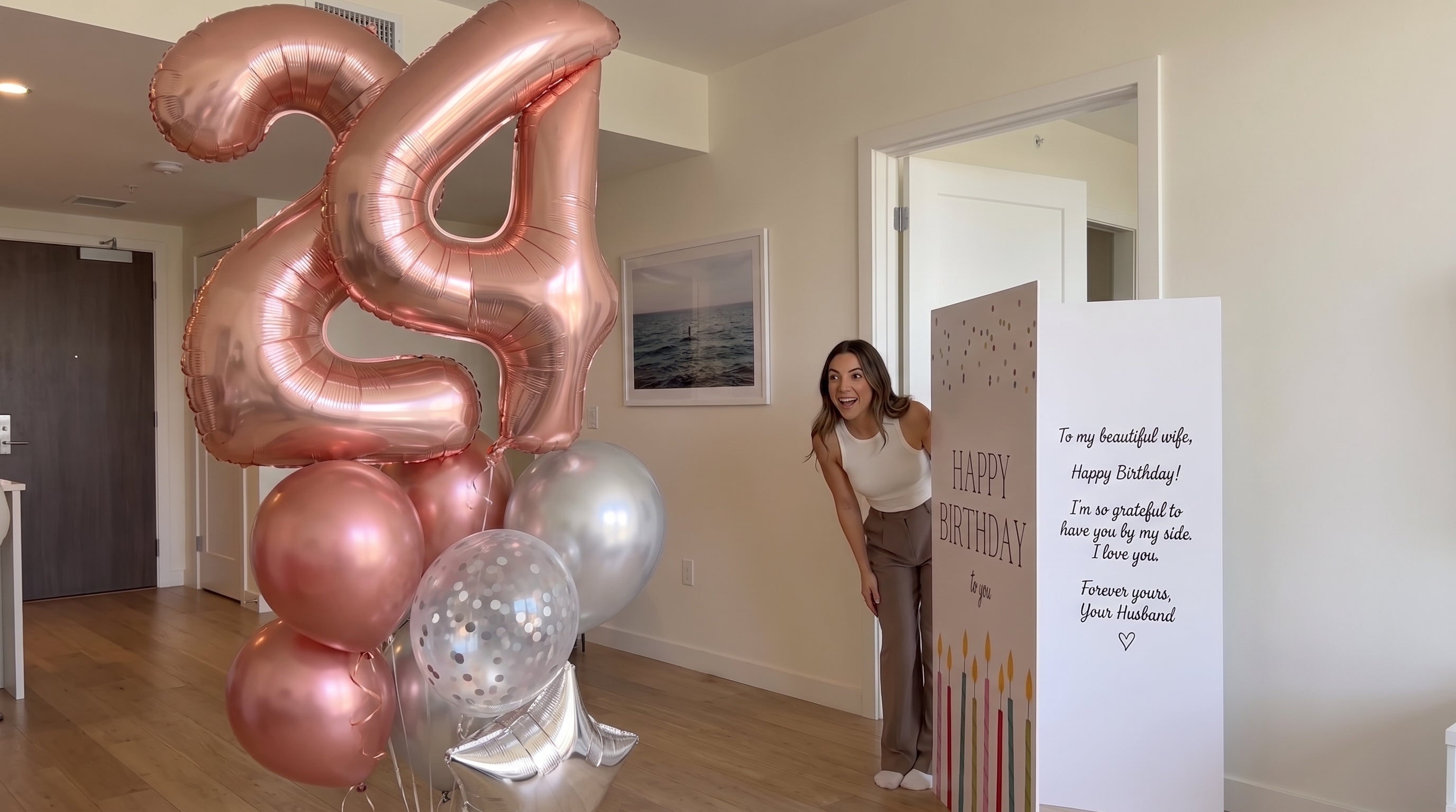 A woman stands in a doorway holding a large white birthday card that reads 'HAPPY BIRTHDAY' with colorful candle illustrations, next to a cluster of metallic rose gold and silver balloons in a home interior.