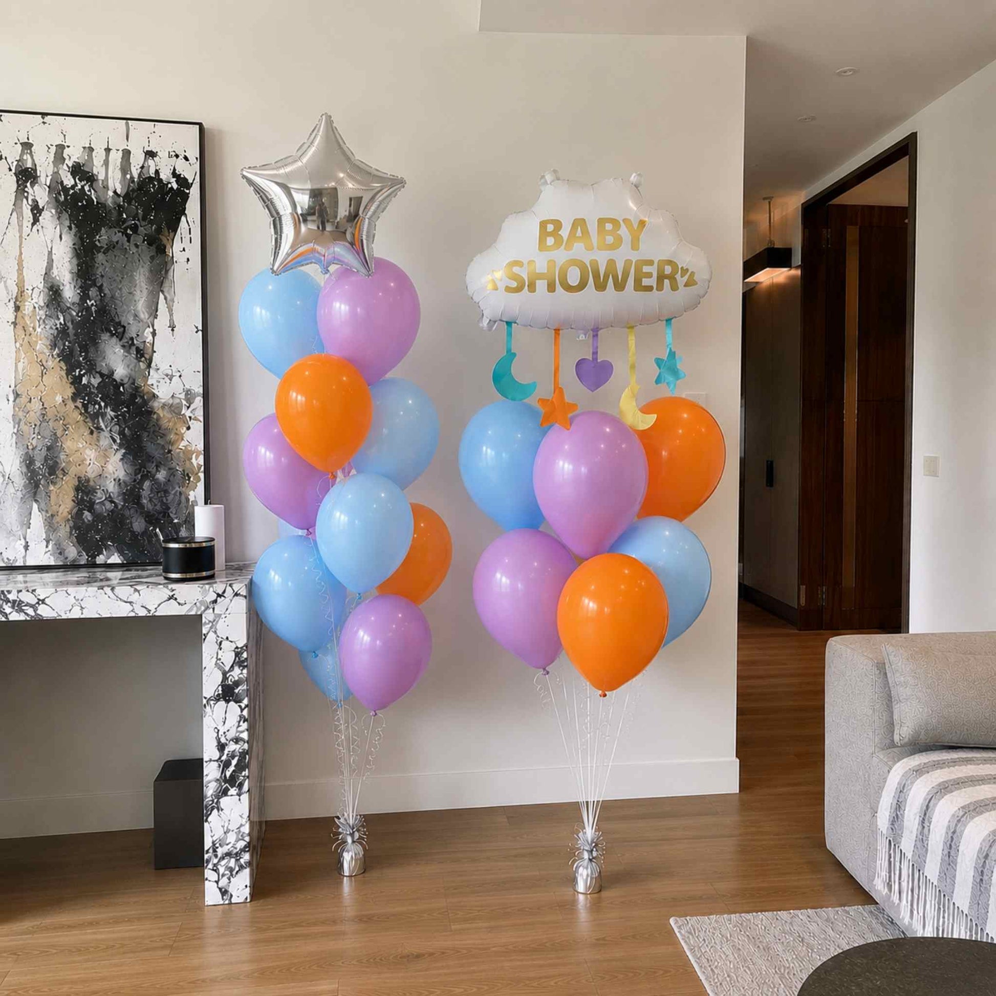 Baby shower balloon bouquet featuring a white Baby Shower cloud foil balloon, silver star foil balloon, and pastel blue, purple, and orange helium latex balloons arranged on silver weights in a modern home.