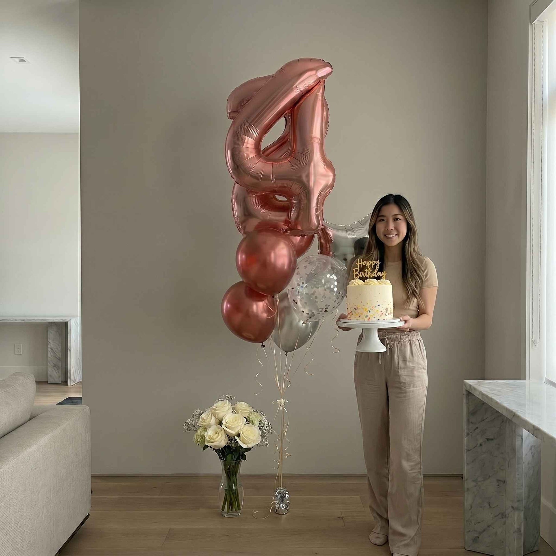Woman holding a cake with large '4' shaped balloons and flowers in a modern living room.
