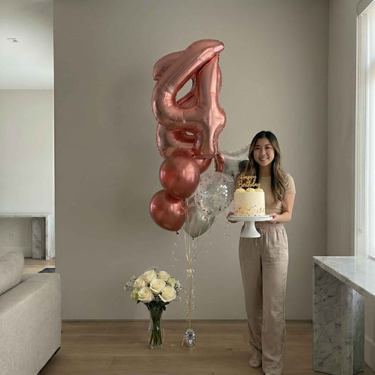 Woman holding a cake with large '4' shaped balloons and flowers in a modern living room.