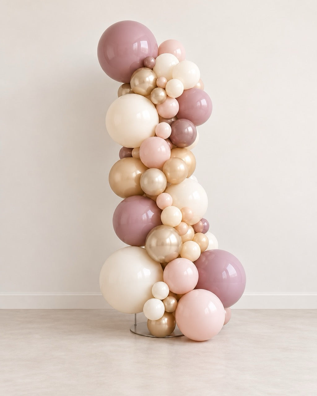 A decorative balloon garland arrangement made of large and small matte and metallic balloons in dusty pink, blush, cream, and champagne gold, displayed on a clear stand against a plain light beige wall and floor.