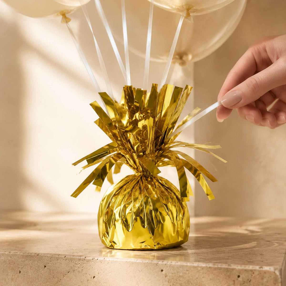 A gold metallic tinsel party decoration with a faceted base and balloon strings, shown on a tabletop with a hand adjusting one of the clear balloons.