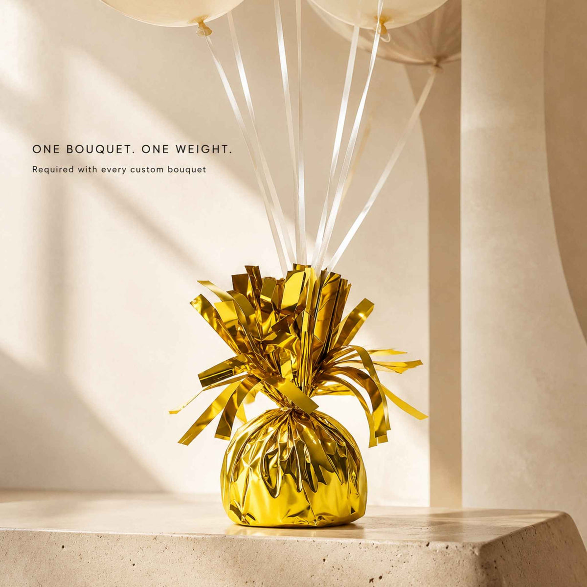 A gold metallic tinsel balloon bouquet decoration sitting on a light stone surface, with white balloons and white ribbons above it against a warm beige architectural background. Text on the left reads: 'ONE BOUQUET.
