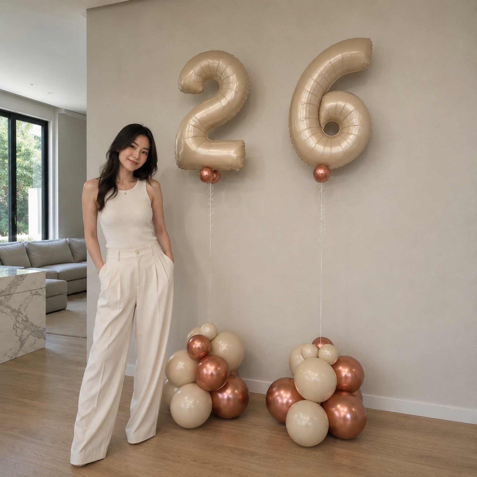 Woman standing next to large metallic number balloons spelling '26' in a modern living room.