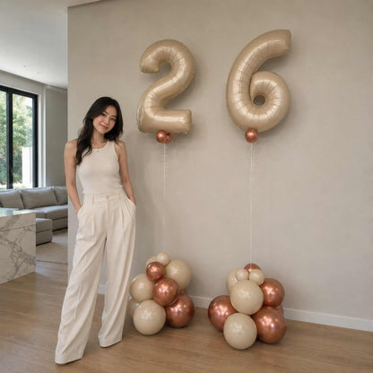 Woman standing next to large metallic number balloons spelling '26' in a modern living room.
