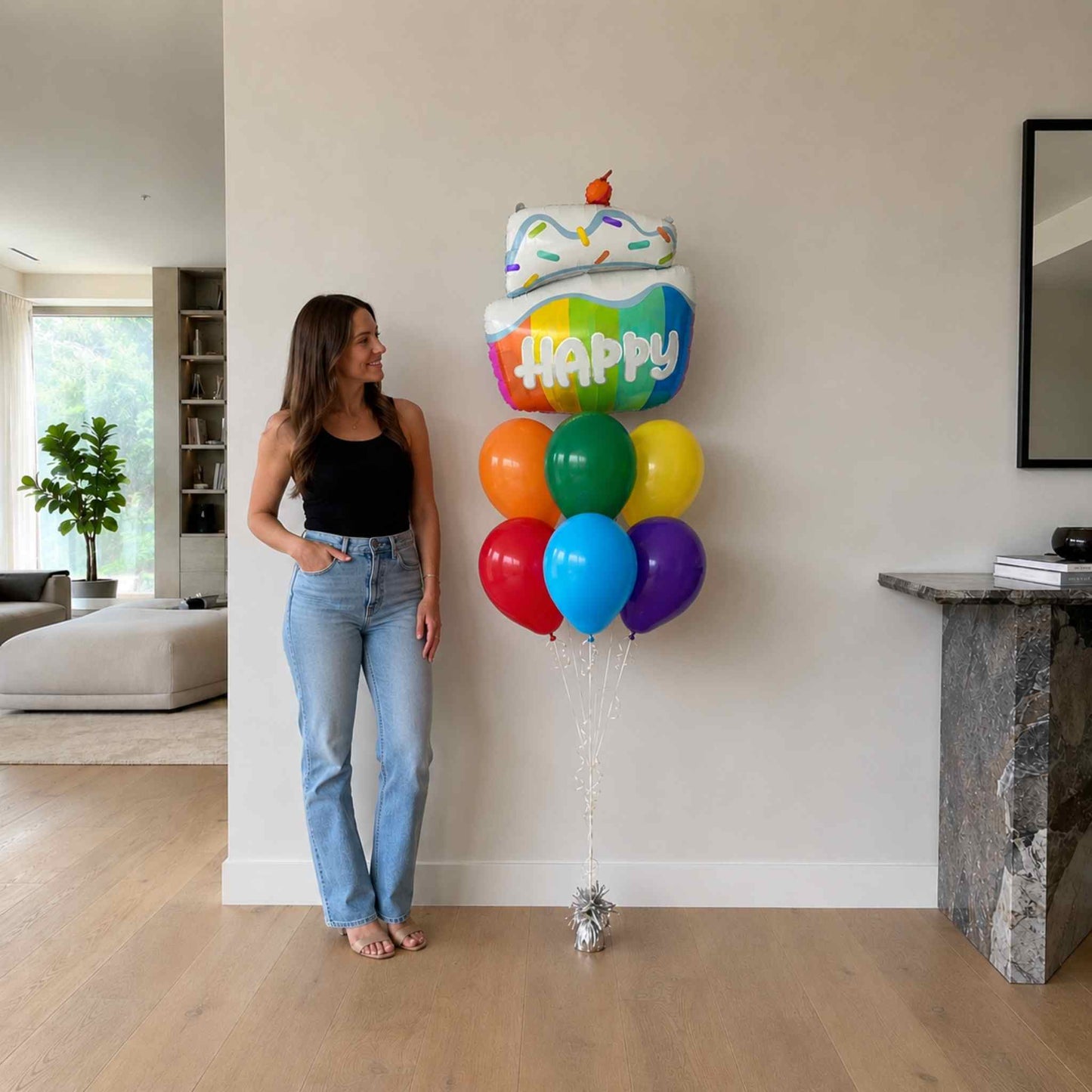 Woman standing next to a large balloon shaped like a birthday cake with colorful balloons in a living room.
