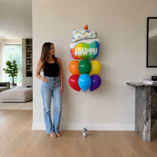 Woman standing next to a large balloon shaped like a birthday cake with colorful balloons in a living room.