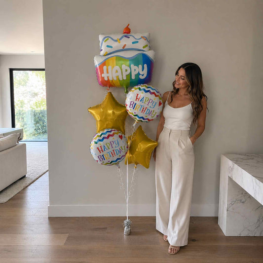 Woman in a white outfit standing next to colorful birthday balloons in a room.