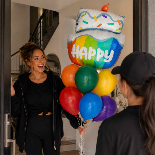 A woman smiling and holding a colorful birthday balloon bouquet with a cake topper and a large rainbow 'HAPPY' balloon, standing in a doorway next to another person wearing a black cap.