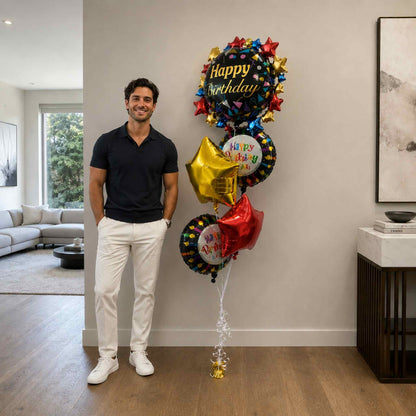 Man standing next to a bouquet of colorful balloons with 'Happy Birthday' text in a living room.