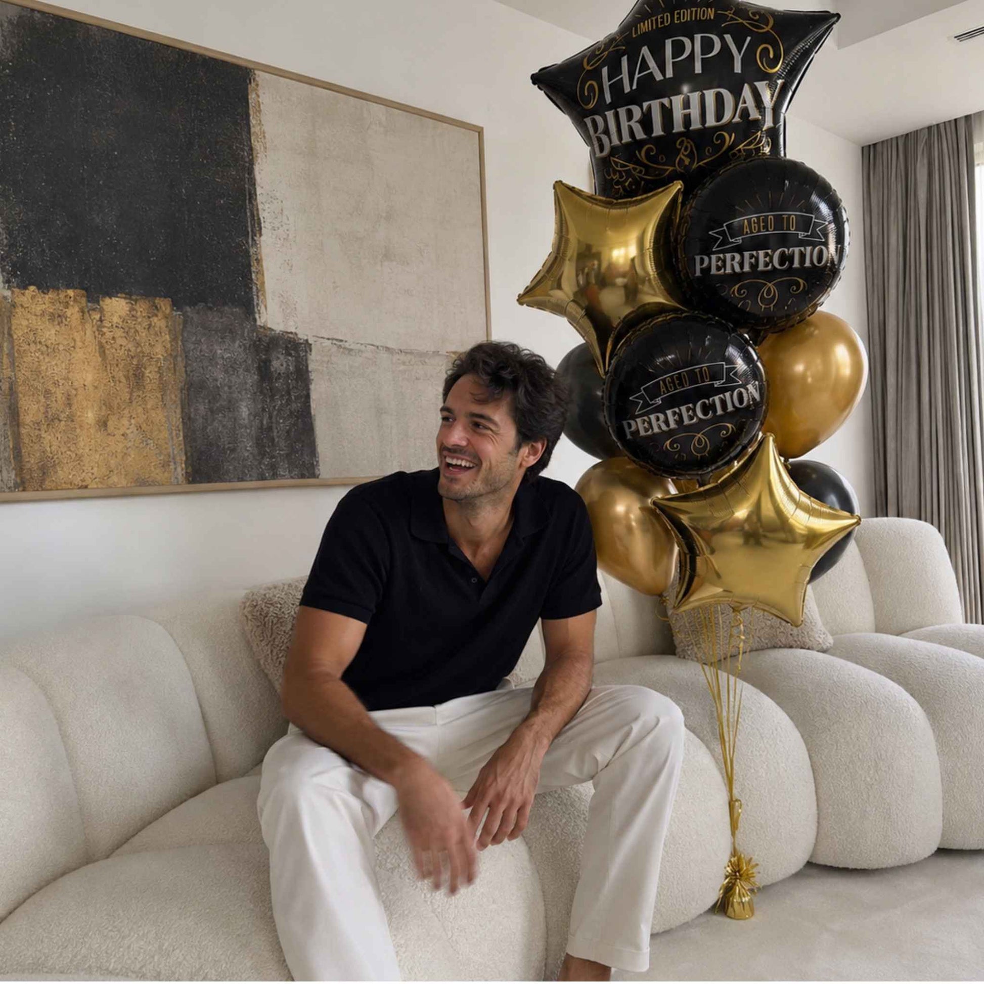 A man in a black polo shirt and white pants sits on a cream-colored boucle sofa next to a cluster of black and gold birthday balloons, including a star-shaped balloon that reads 'HAPPY BIRTHDAY' and round balloons that say 'PERFECTION'.