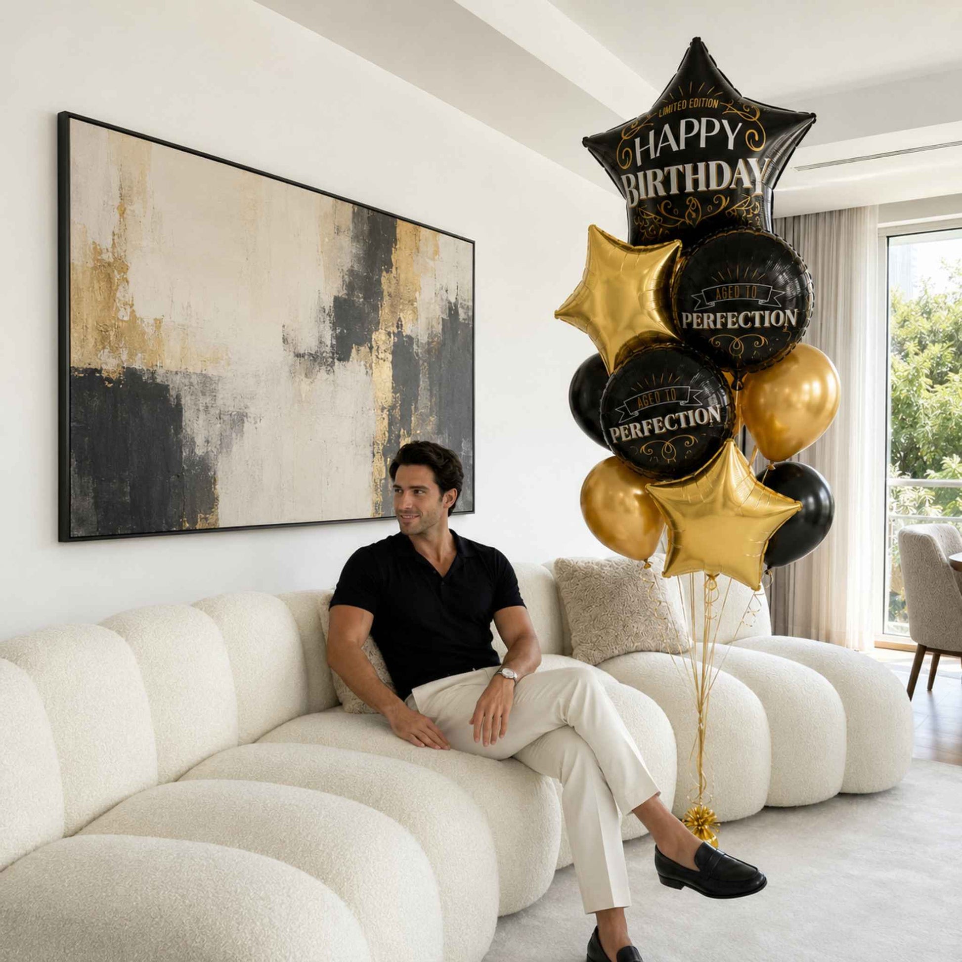A man sits on a modern curved white sofa in a bright living room, next to a large balloon bouquet featuring gold and black balloons with 'HAPPY BIRTHDAY' and 'PERFECTION' text.
