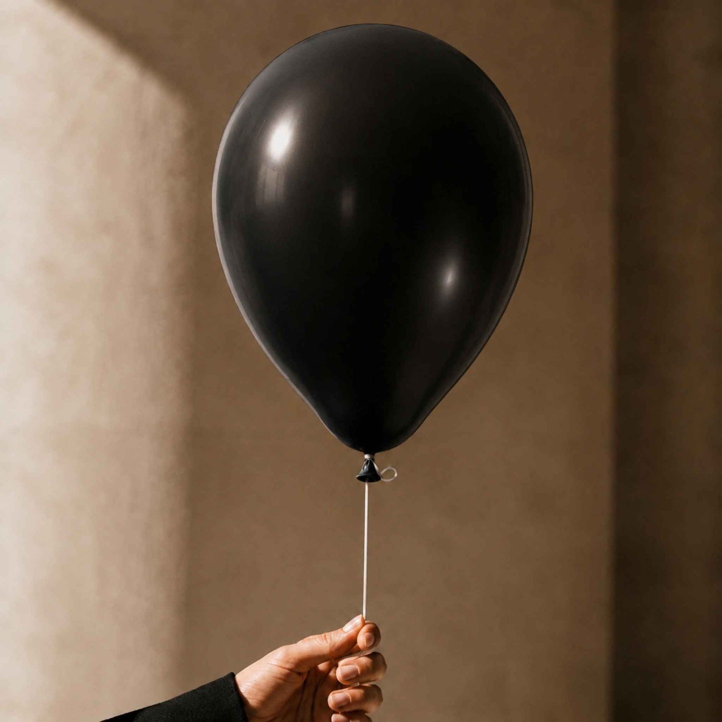 A matte black latex balloon held by a hand against a warm beige textured wall.