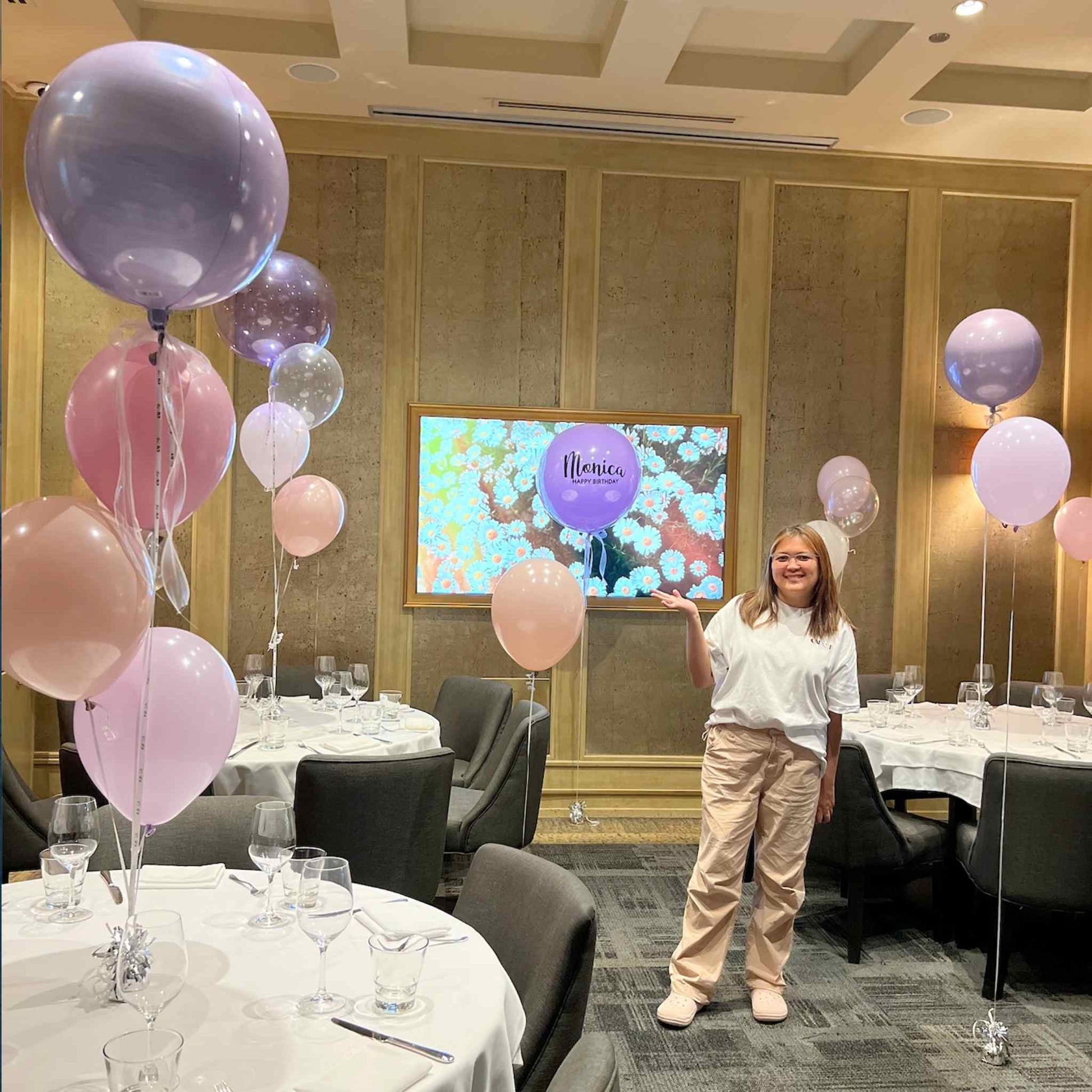 Person standing in a decorated room with balloons and tables, smiling.
