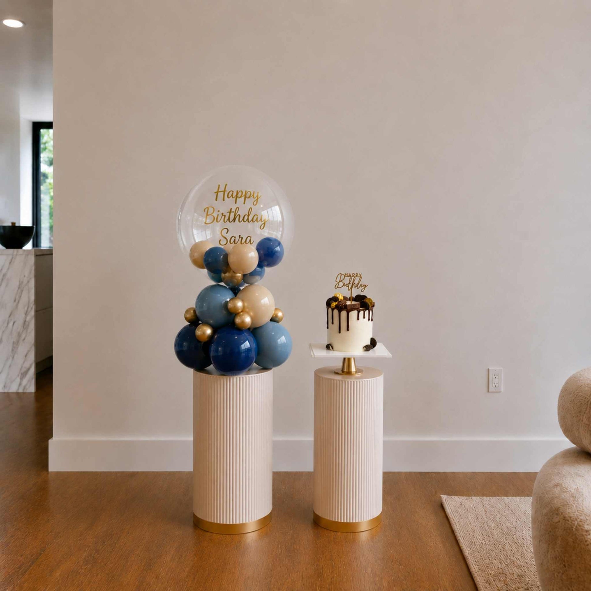 Decorative balloon display with 'Happy Birthday Sara' on a stand next to a small cake on a matching stand.