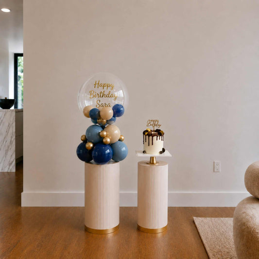 Decorative balloon display with 'Happy Birthday Sara' on a stand next to a small cake on a matching stand.