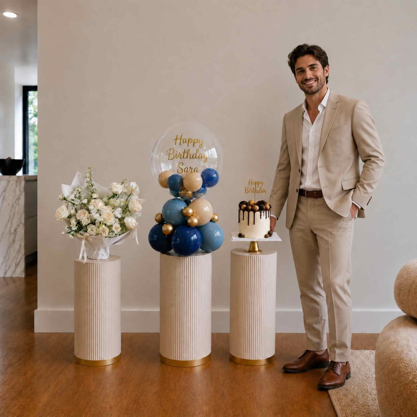 Man in a beige suit standing next to a birthday setup with balloons, cake, and flowers.