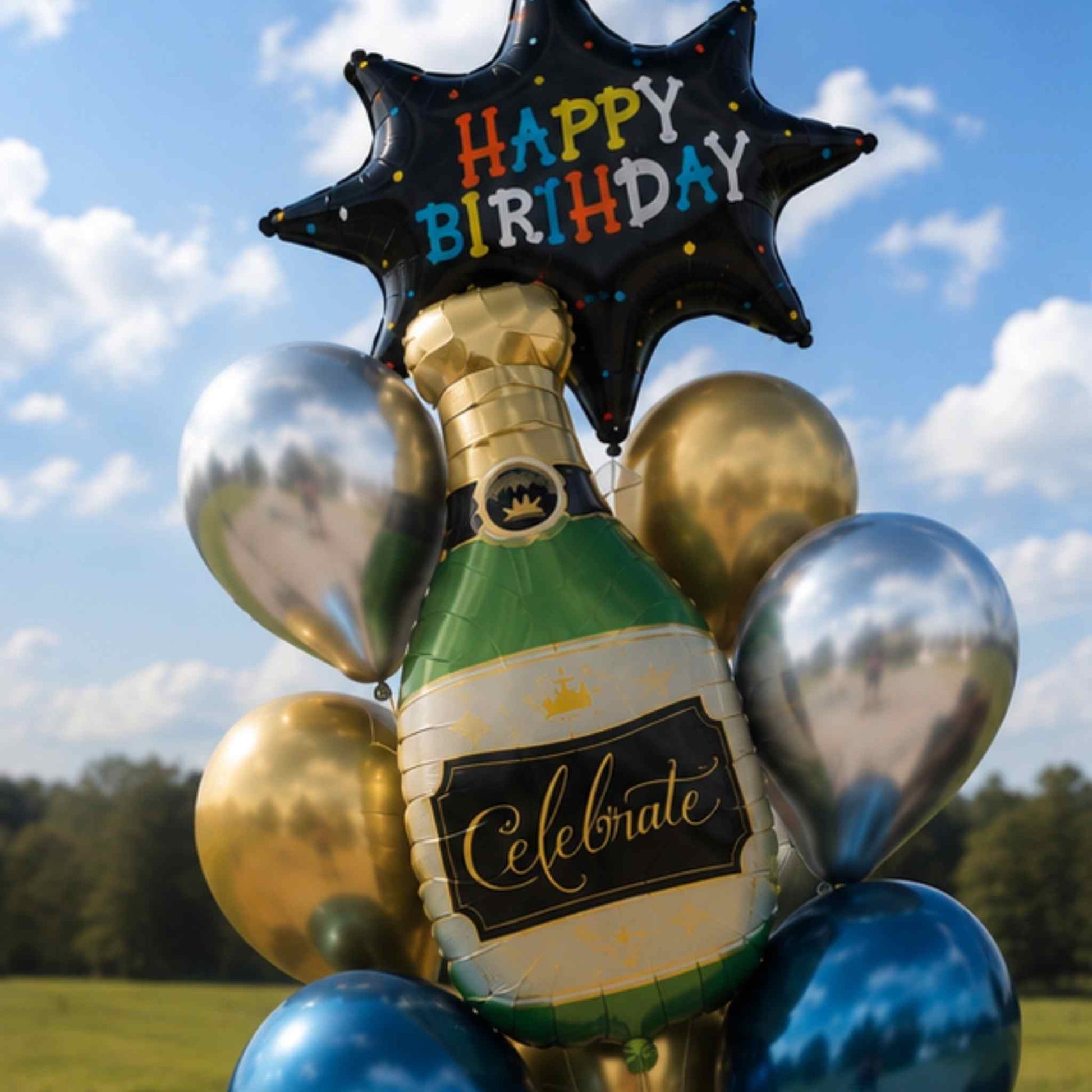 A birthday balloon bouquet featuring a champagne bottle-shaped foil balloon with a black label reading 'Celebrate,' surrounded by metallic silver, gold, and blue balloons, topped with a black star-shaped balloon that says 'HAPPY BIRTHDAY.' The scene is photographed outdoors against a blue sky with clouds and trees in the distance.
