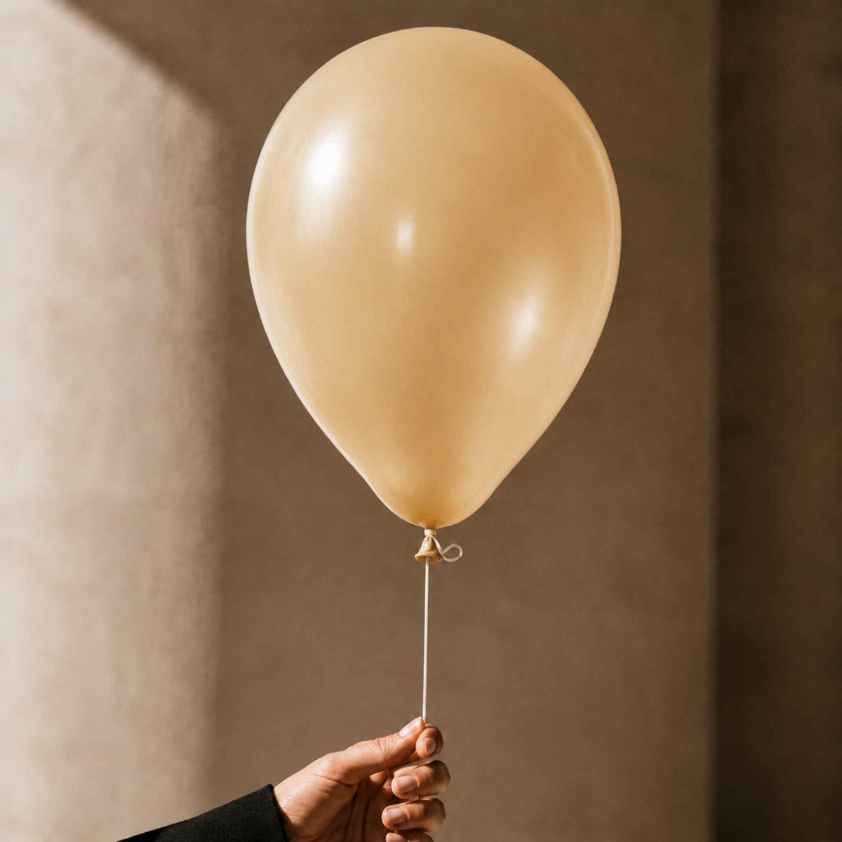 A person's hand holding the string of a single matte champagne-gold balloon against a warm, textured beige background.