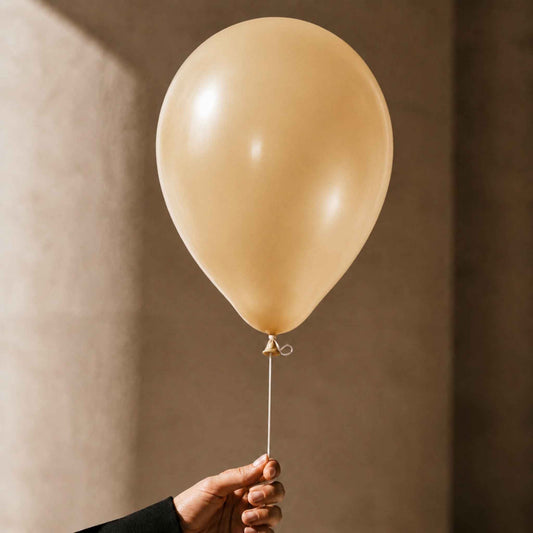 A person's hand holding the string of a single matte champagne-gold balloon against a warm, textured beige background.