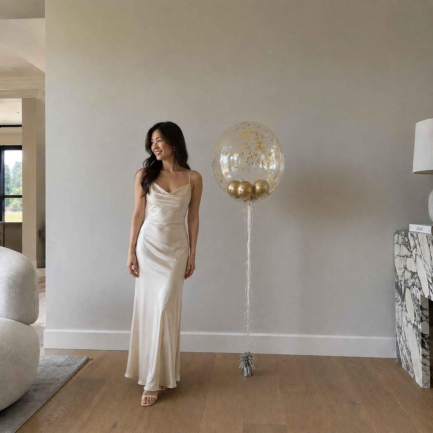 Woman in a long, light-colored dress standing next to a large decorative balloon in a modern living room.