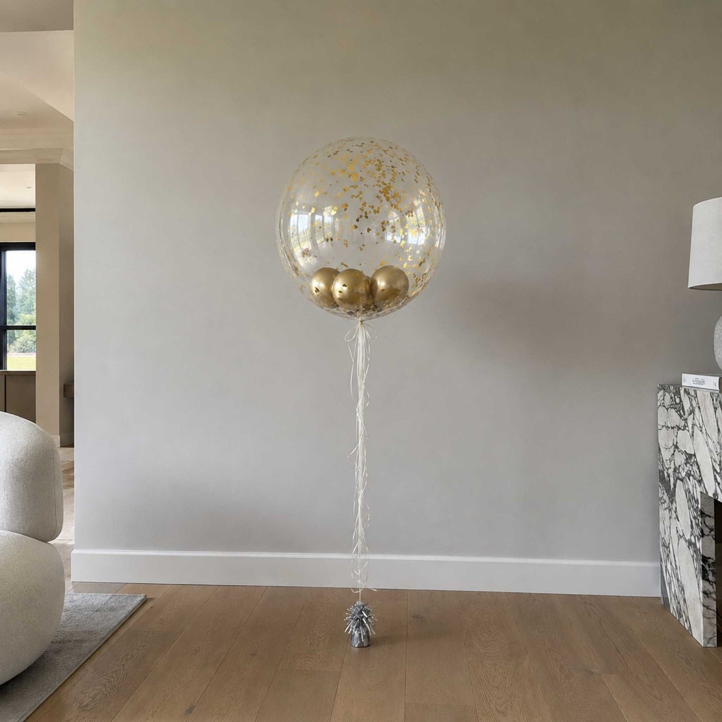 Large transparent balloon with gold confetti and balloons inside, tied to a small silver object on a wooden floor.