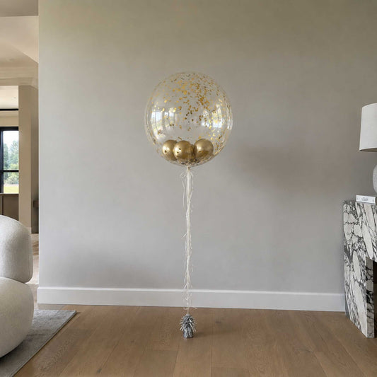 Large transparent balloon with gold confetti and balloons inside, tied to a small silver object on a wooden floor.