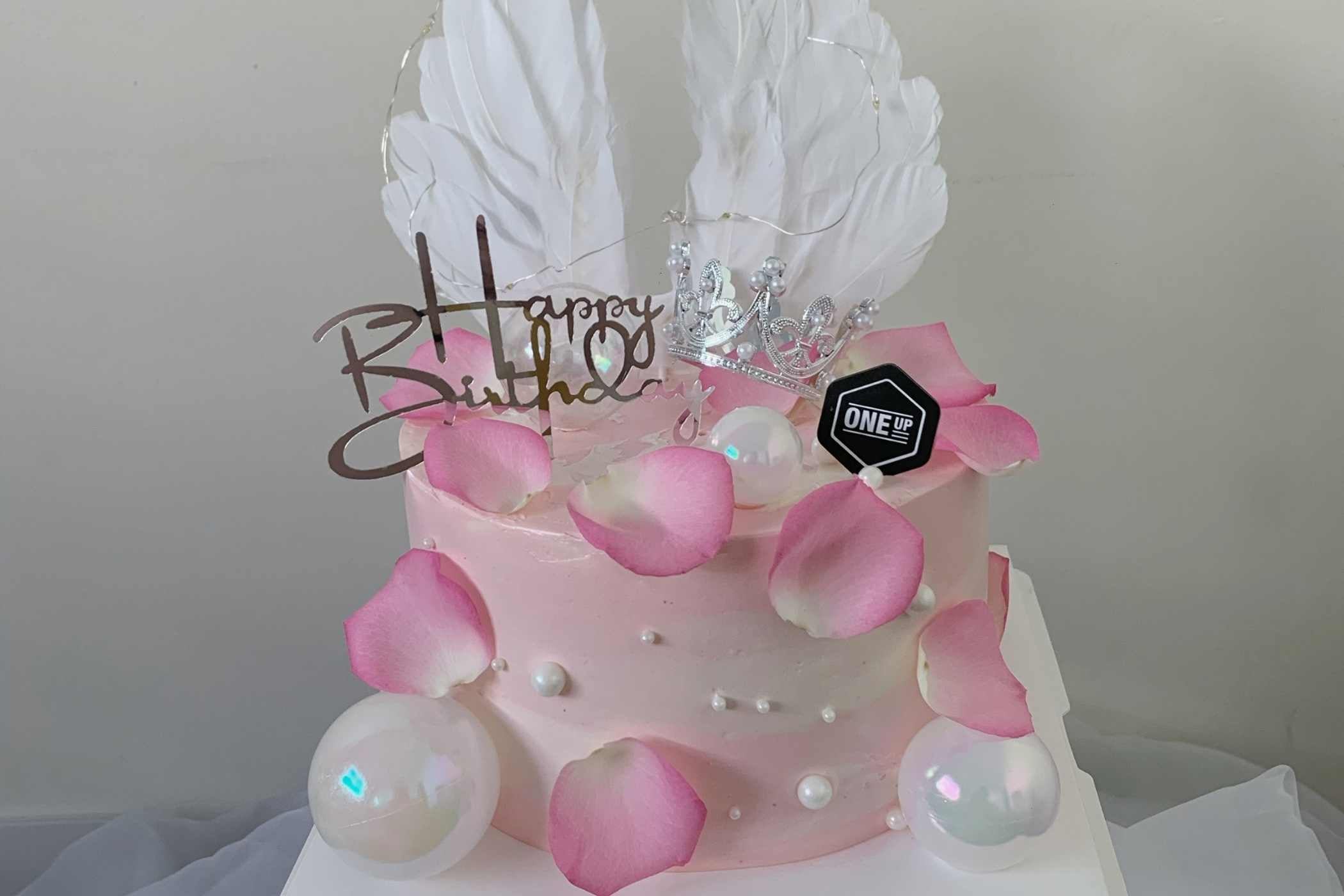 Pink birthday cake with white flowers and 'Happy Birthday' topper on a plain background
