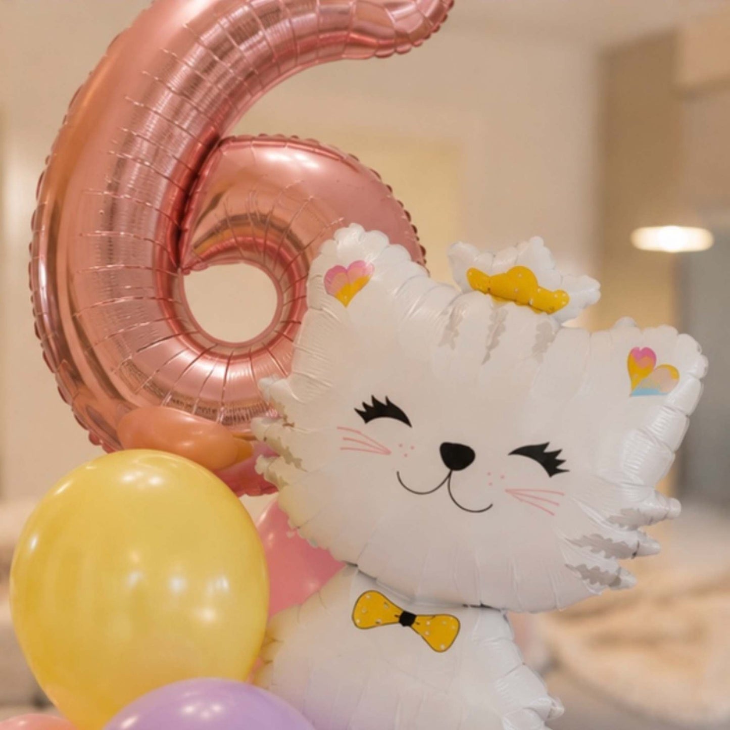 A white cartoon cat foil balloon with a yellow bow tie and small heart accents, posed with a large metallic pink number 6 balloon and other pastel balloons in an indoor setting.