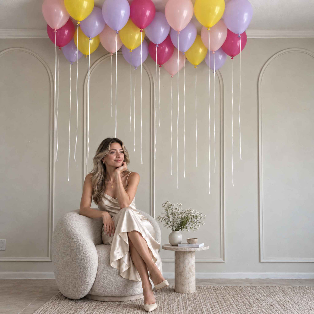 A woman in a champagne satin dress sits in a textured cream boucle armchair, with a cluster of pastel pink, purple, yellow, and red balloons floating above her in a decorated room with cream paneled walls.